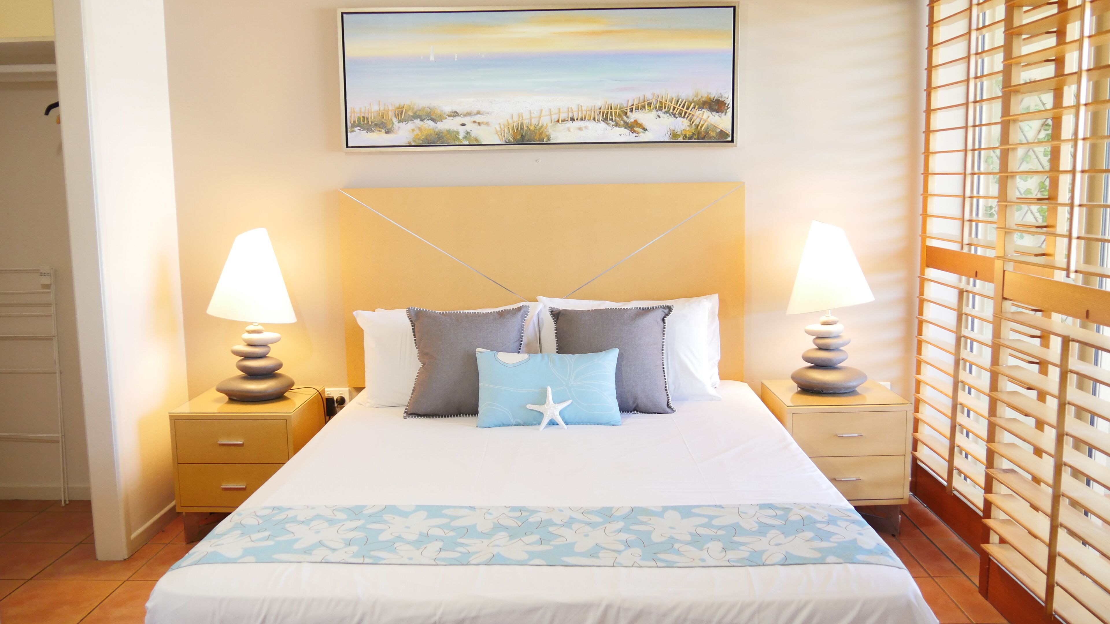 A neatly made bed with a floral bedspread, two bedside lamps, and a painting of a beach scene above it.