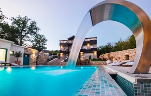 Ett modernt poolområde med ett tydligt vattenelement, stenmurar och en tvåvåningsbyggnad med balkonger.