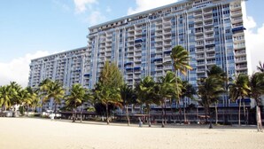 Un hotel grande con varios pisos y balcones, ubicado en una playa de arena con palmeras.