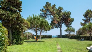 A well-maintained grassy area with a playground, large trees, and a clear view of the sea.