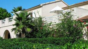 A building with a white facade, a red-tiled roof, and a white umbrella. There are green bushes and a palm tree in the foreground.