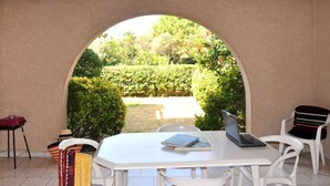 A white table with a laptop and a book, surrounded by white chairs, under a semi-circular archway with a view of a garden.