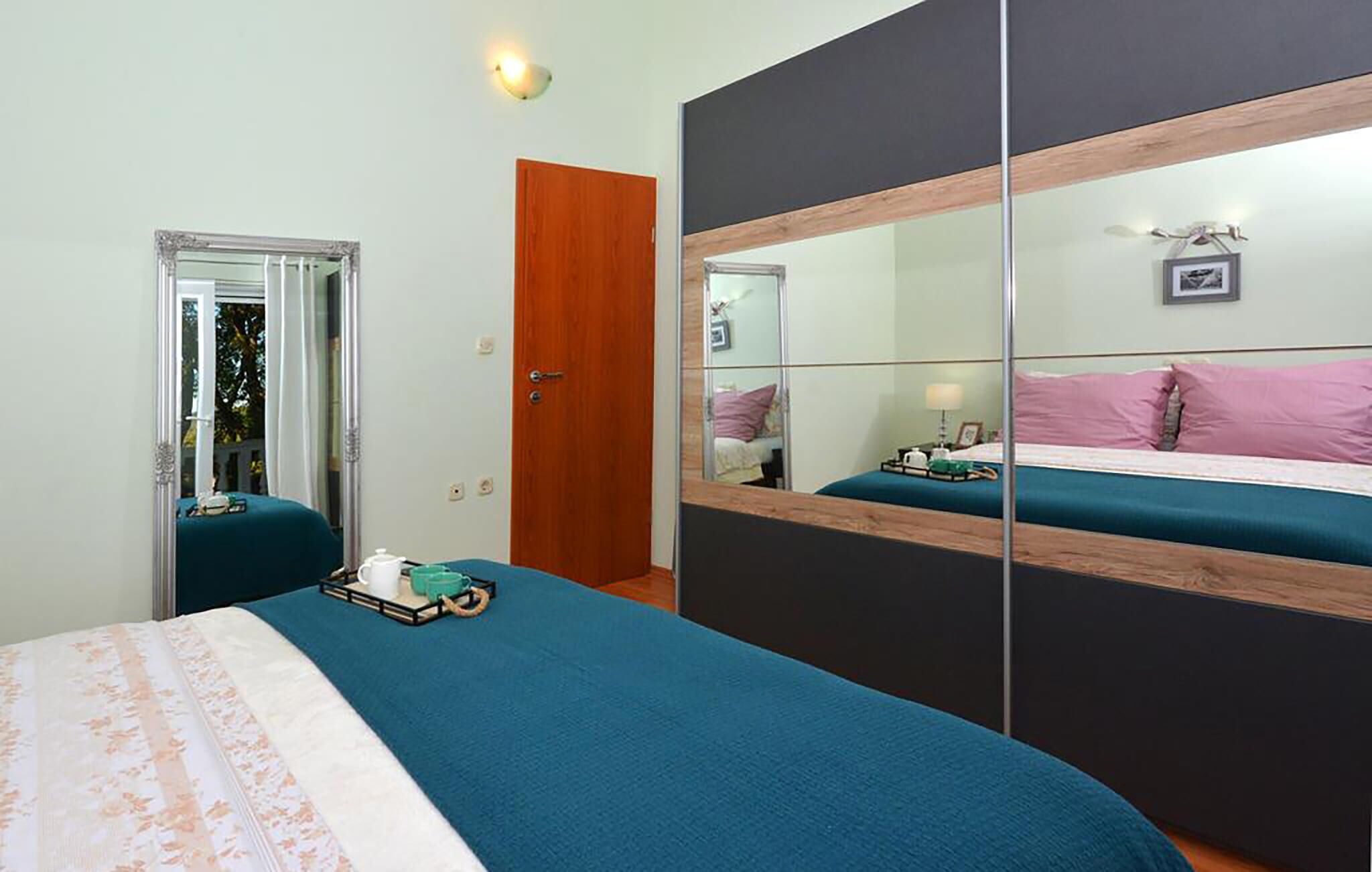 Un dormitorio con cama, armario con espejos, puerta y mesita de noche con lámpara.