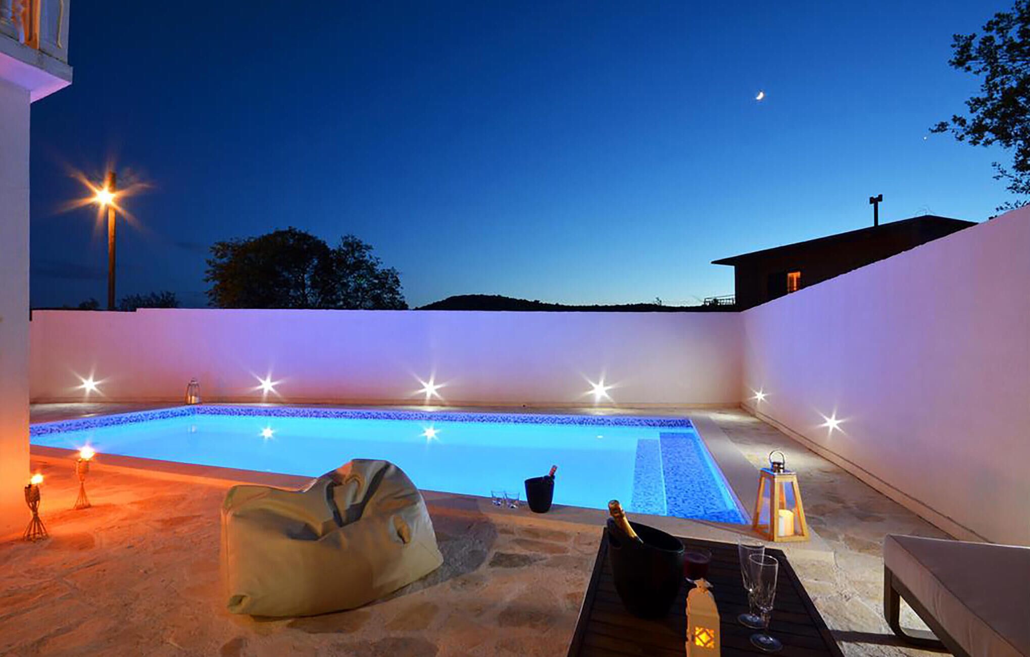 Uma área de piscina ao ar livre com um deck de pedra, uma espreguiçadeira e uma mesa com bebidas e um candeeiro.