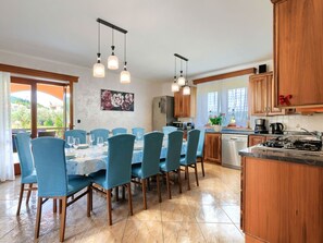 Uma sala de jantar com mesa posta para seis pessoas, cercada por cadeiras azuis, uma cozinha com armários de madeira e vista para o verde lá fora.