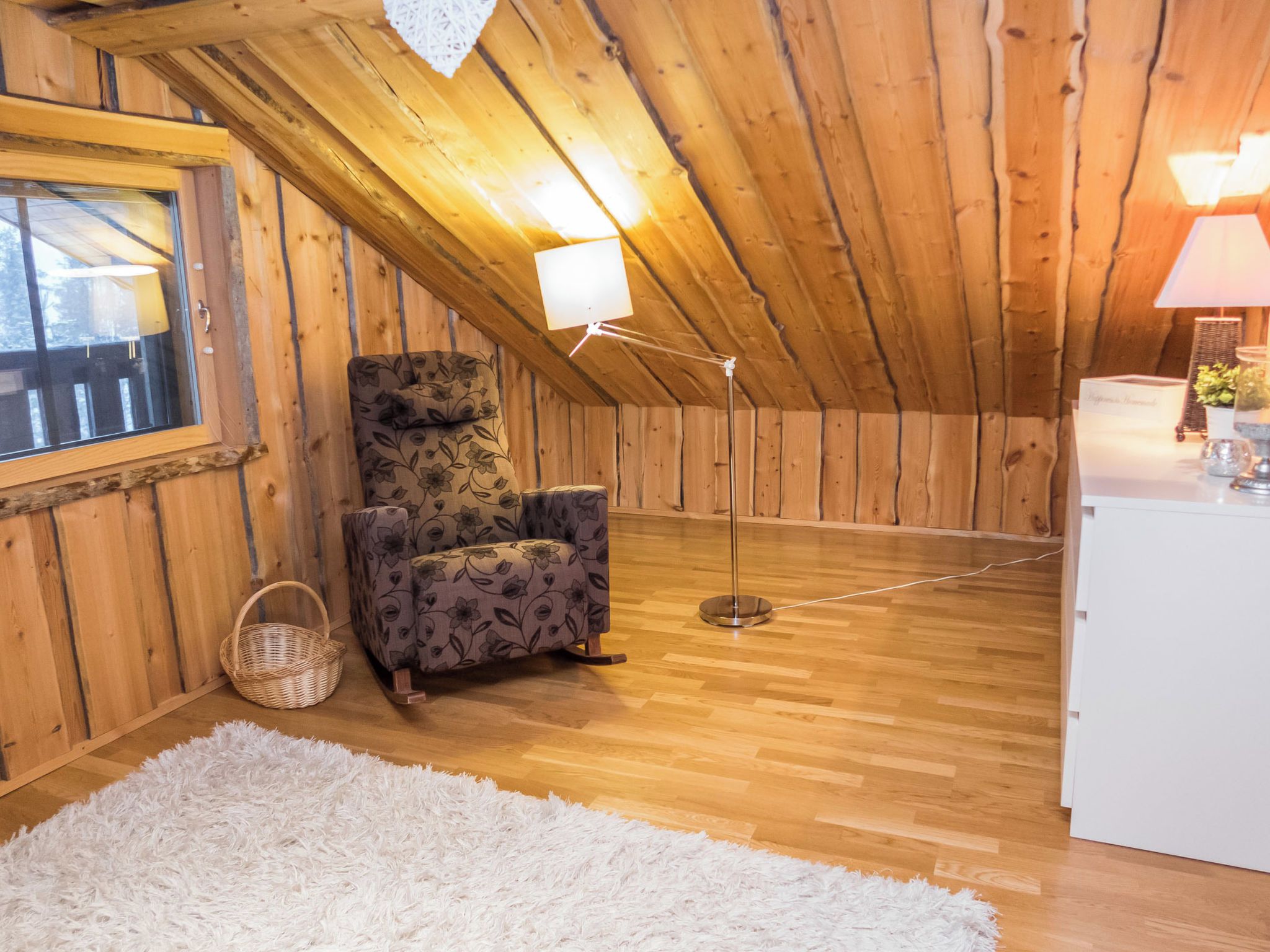 A cozy attic room with wooden walls, a floral-patterned chair, a white rug, and a floor lamp.