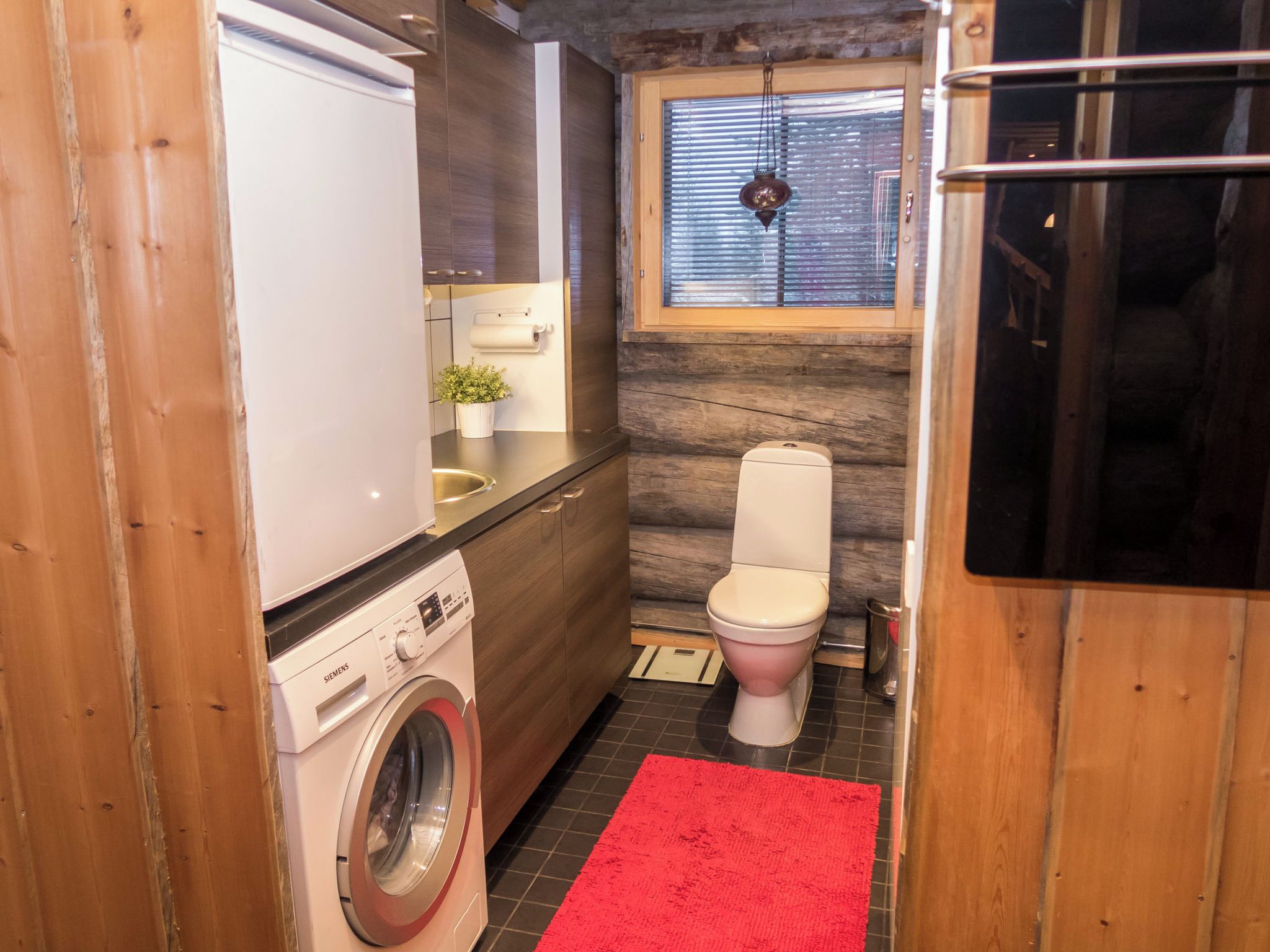 A compact bathroom with a washing machine, toilet, and a small window with blinds.