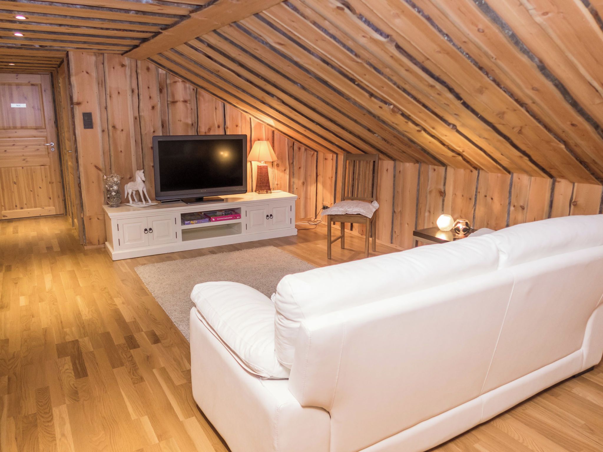 A cozy attic room with a white sofa, a TV on a wooden cabinet, and a small table with a lamp.