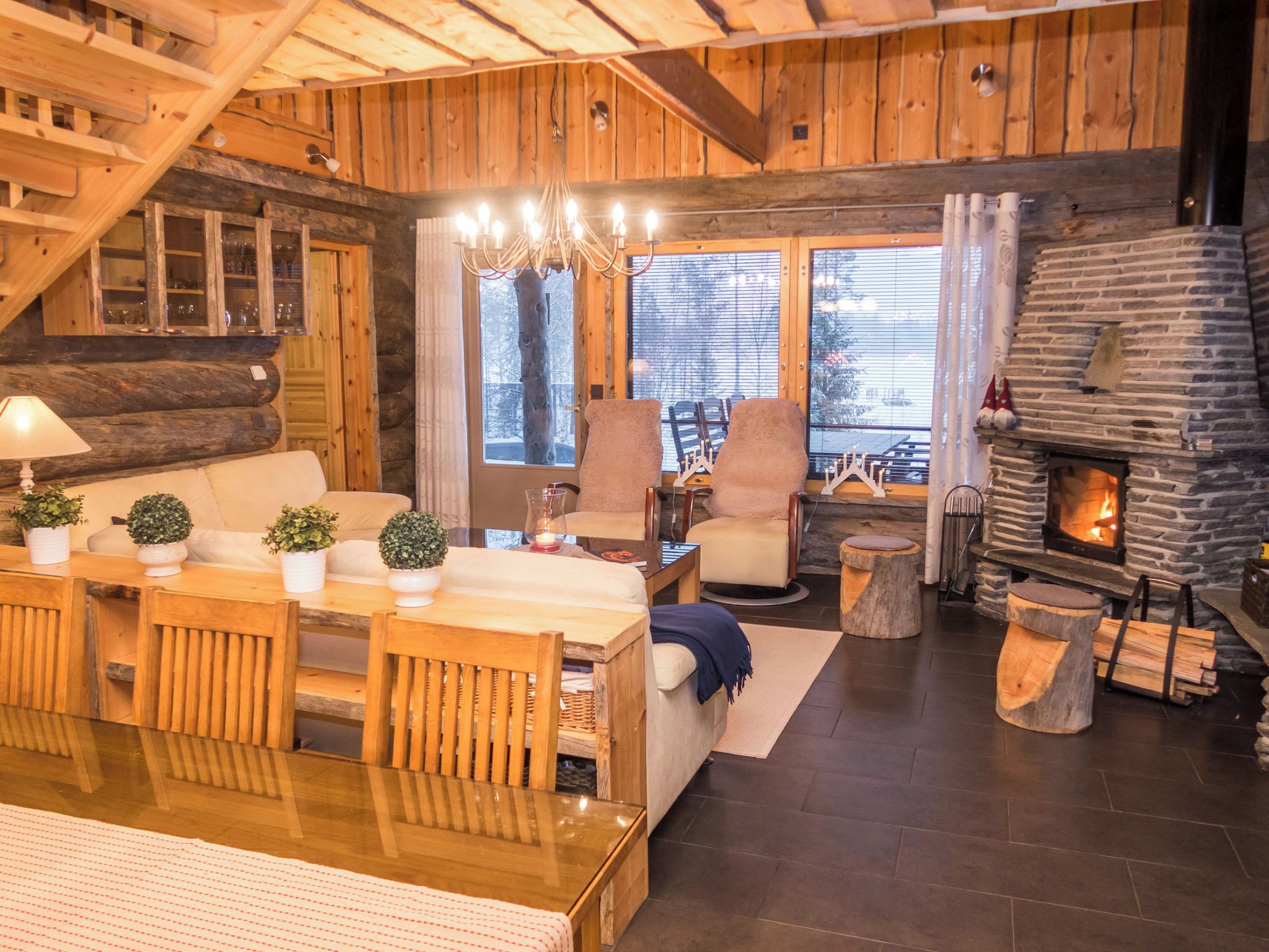 A cozy log cabin interior with a stone fireplace, wooden furniture, and a view of the snowy outdoors.