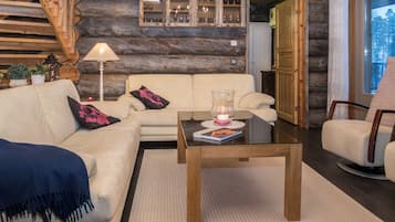 A cozy living room with a sofa, armchair, and coffee table. There is a fireplace, a lamp, and a bookshelf with books and decorative items.