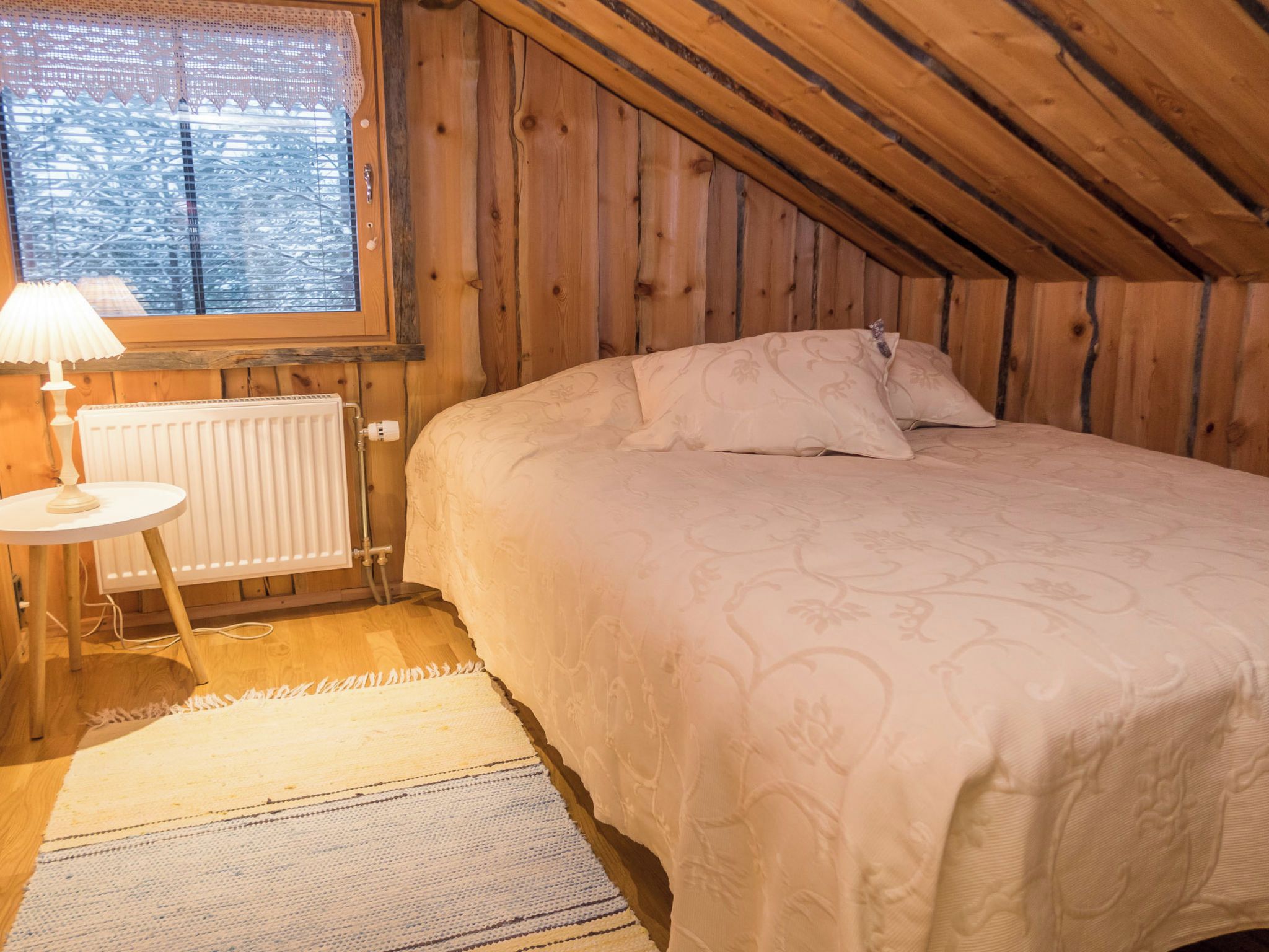 Una camera da letto con un letto, un comodino, una lampada, un termosifone e una finestra vista alberi.