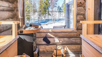 A log cabin interior with a large window, a wood stove, and a sauna bench.
