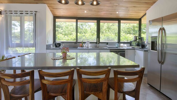 A modern kitchen with a large island, wooden chairs, and a stainless steel refrigerator.