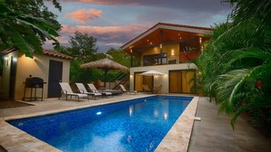 A pool with lounge chairs, a barbecue, and a thatched-roof structure.