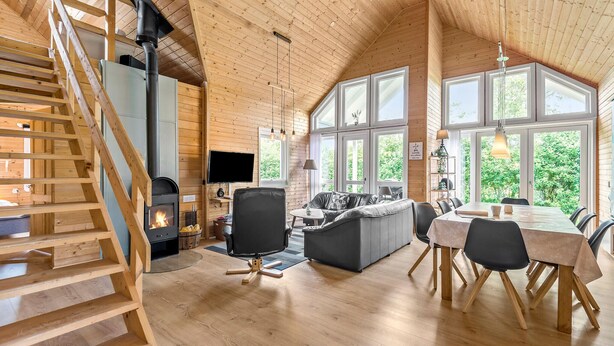 Ruim interieur van een blokhut met een houten trap, een open haard, een bank, een eettafel en grote ramen met uitzicht op groen.