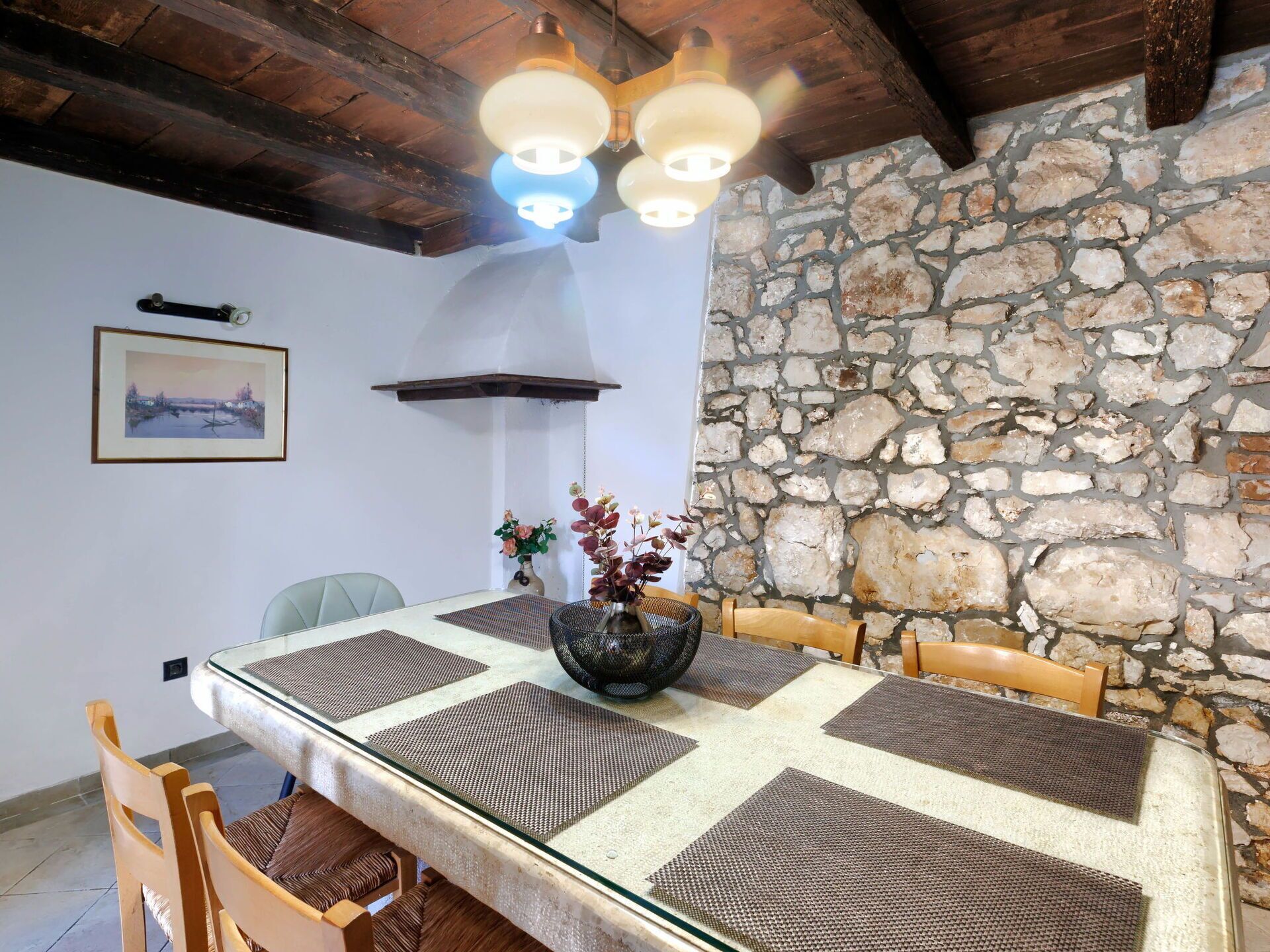 Un comedor con pared de piedra, vigas de madera en el techo y una mesa con individuales y un jarrón de flores.