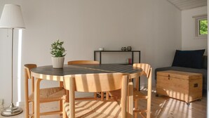 Un comedor con una mesa redonda, cuatro sillas, una planta en maceta, una lámpara de pie, una mesita con adornos y un baúl.