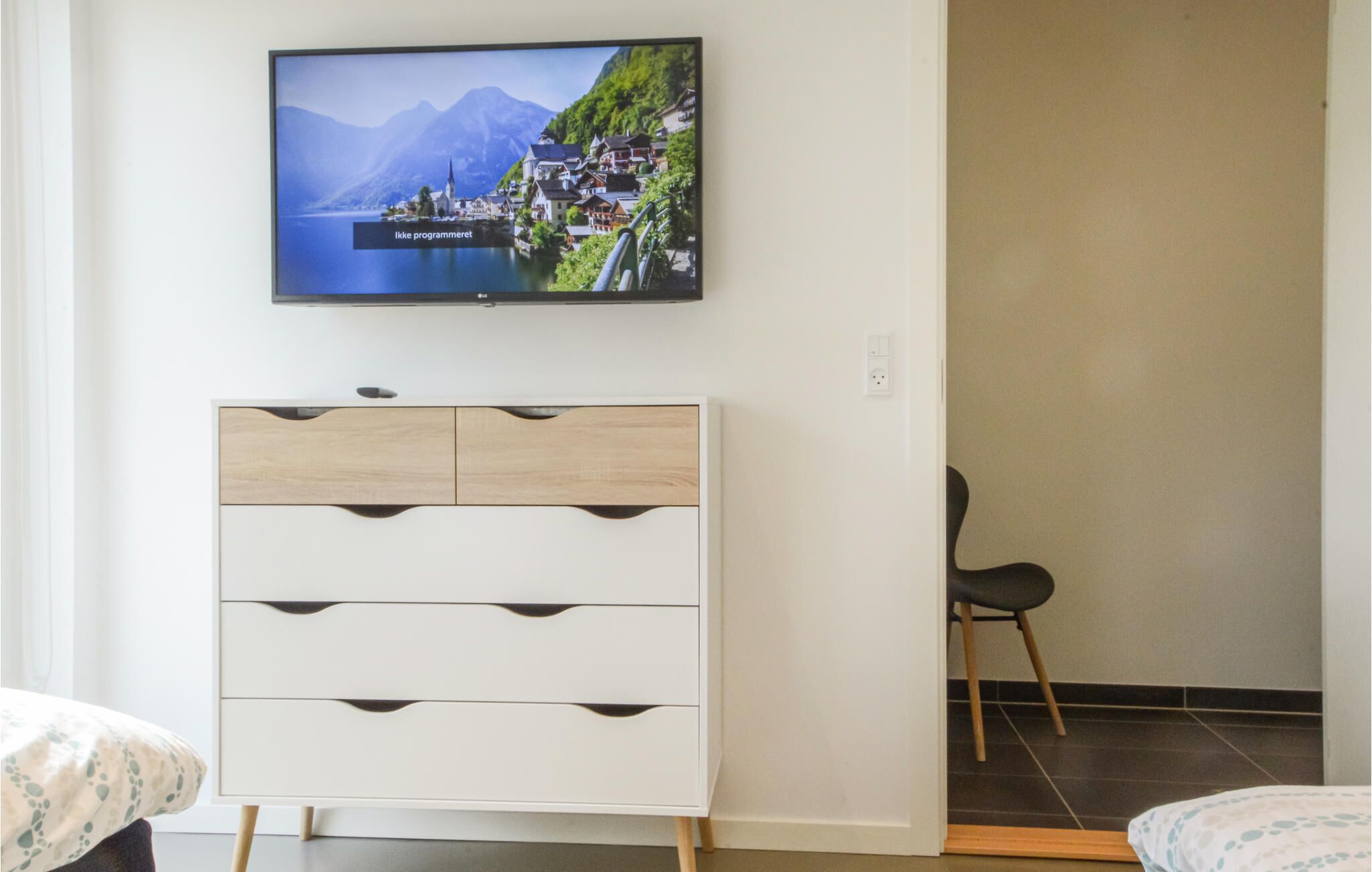 A bedroom with a TV mounted on the wall displaying a scenic image, a white dresser with multiple drawers, a chair, and a bed with a patterned bedspread.