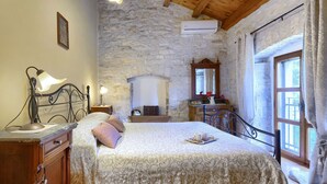 A bedroom with a stone wall, a wooden ceiling, a bed with a floral bedspread, a nightstand with a lamp, a dresser, and a window with curtains.