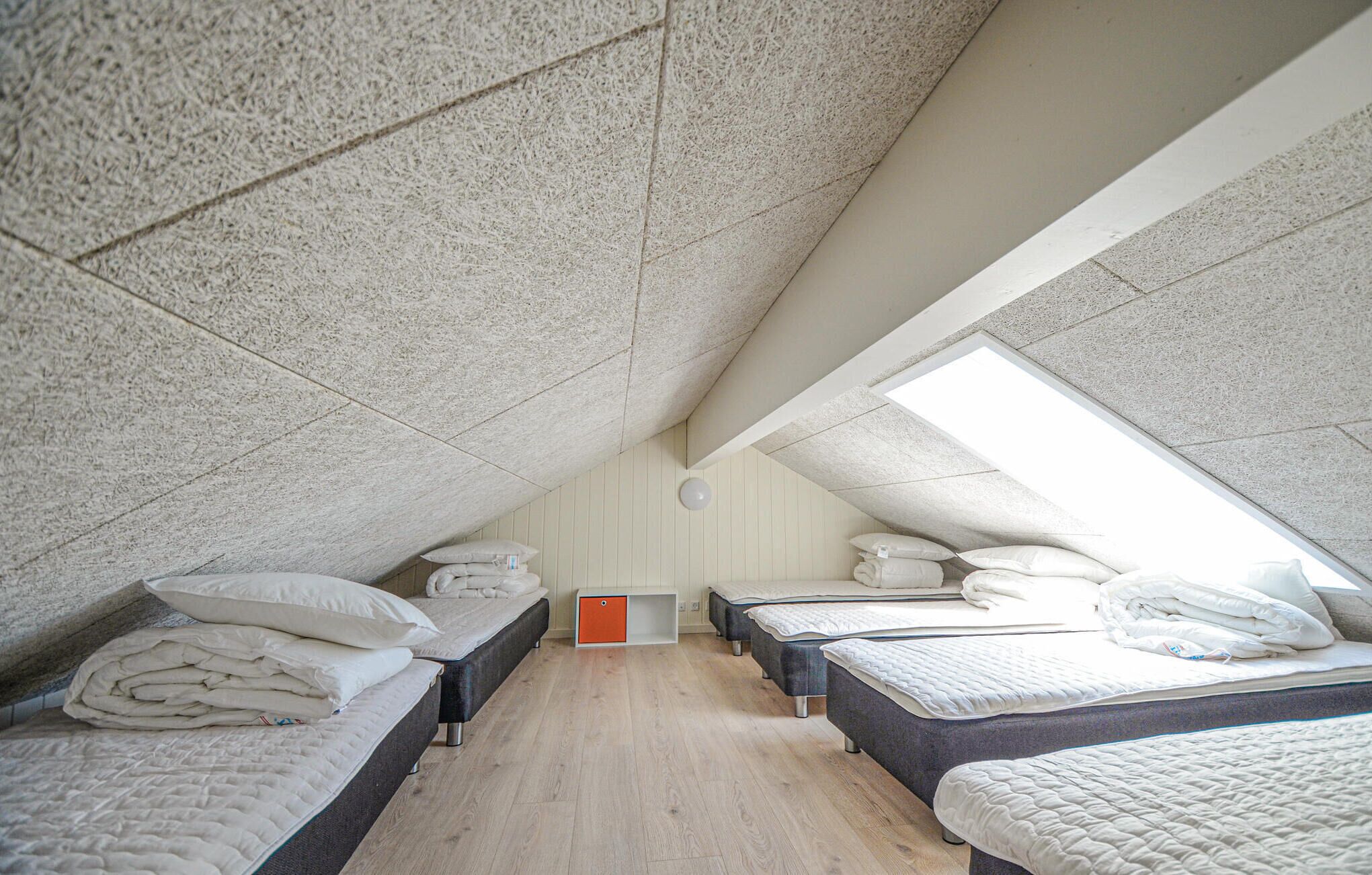 A dormitory-style room with four beds arranged in a row, a wooden floor, and a sloped ceiling with skylights.