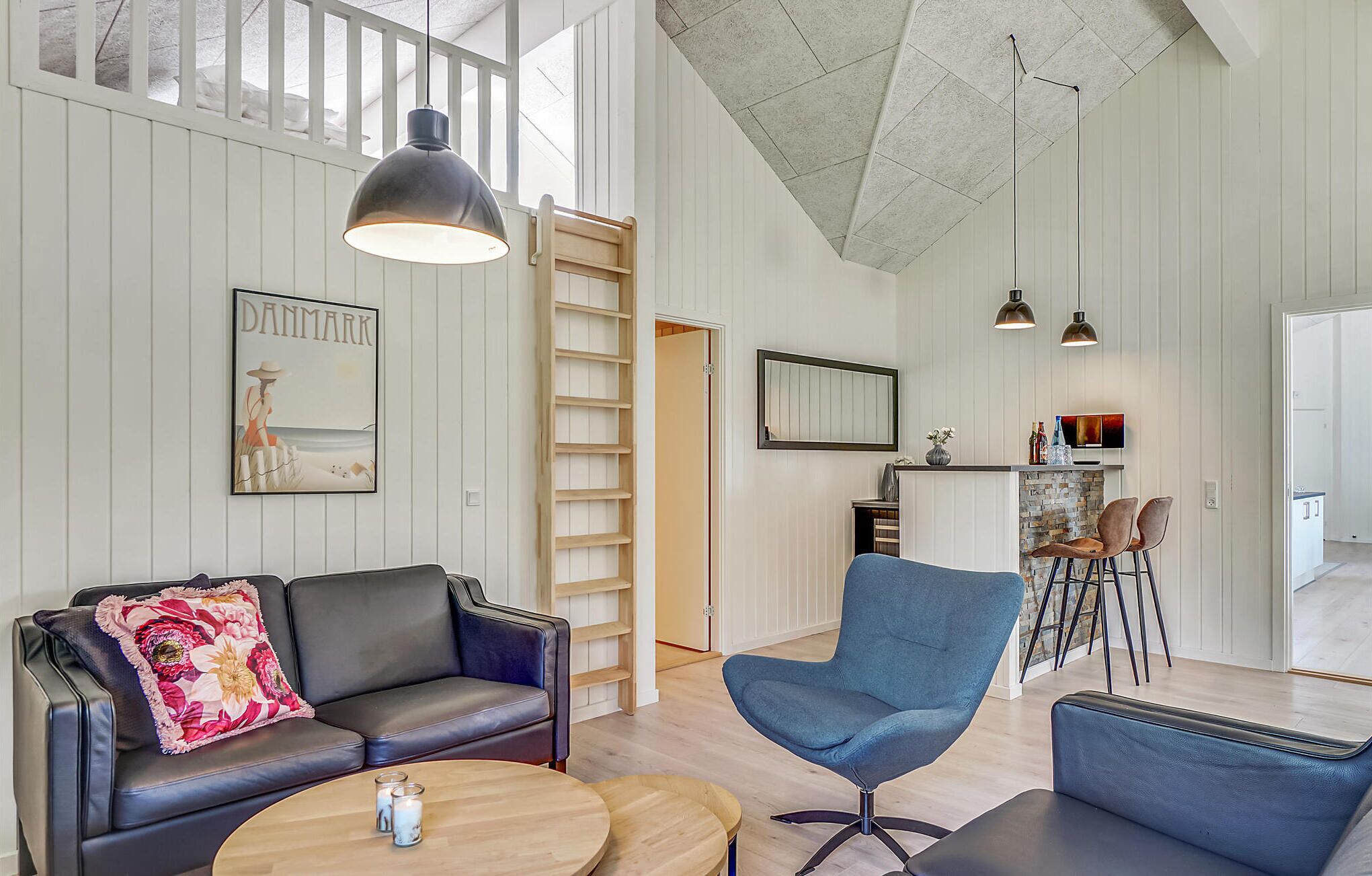 A spacious living area with a black leather sofa, a blue armchair, and a wooden coffee table. A ladder leads to a lofted space, and a television is mounted on the wall.