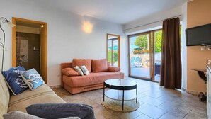 Un salon comprenant un canapé, une table basse et un téléviseur. Une porte-fenêtre coulissante donne accès à un espace extérieur.