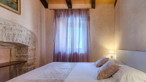 Une chambre avec une cheminée en pierre, un lit recouvert de draps blancs et une fenêtre ornée de rideaux légers.