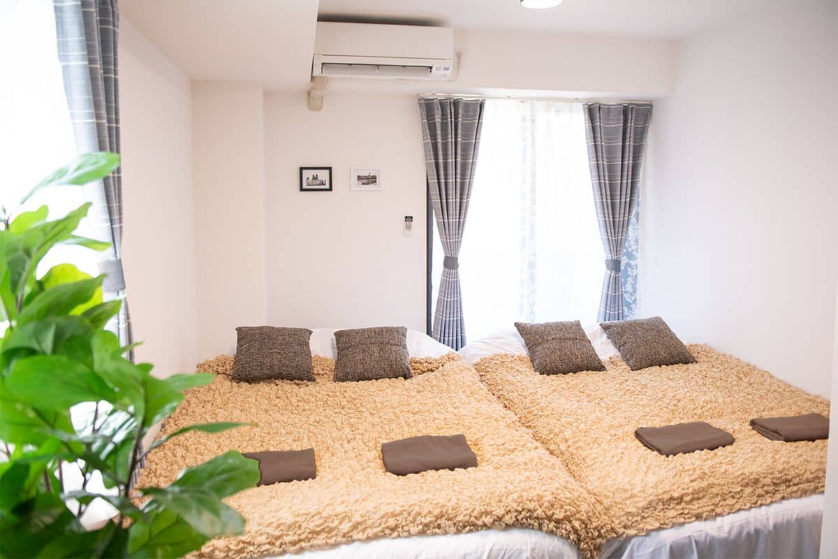 A room with a large bed covered in a textured blanket, two air conditioning units, a window with curtains, and a small framed picture on the wall.
