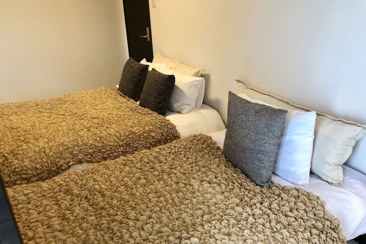 A hotel room with two beds, each with a textured beige blanket and pillows.