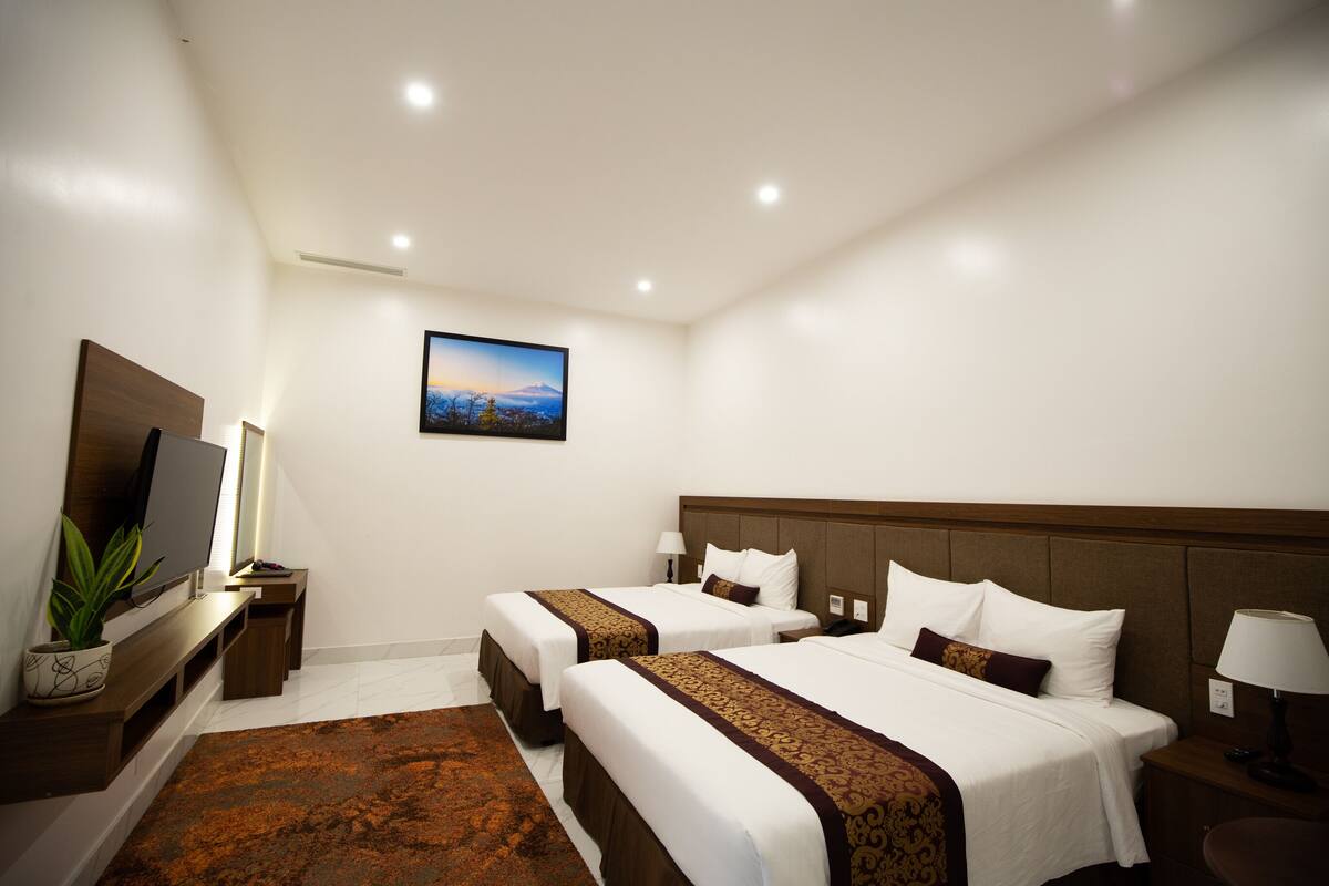 A hotel room with two beds, a TV, a plant, and a painting on the wall.