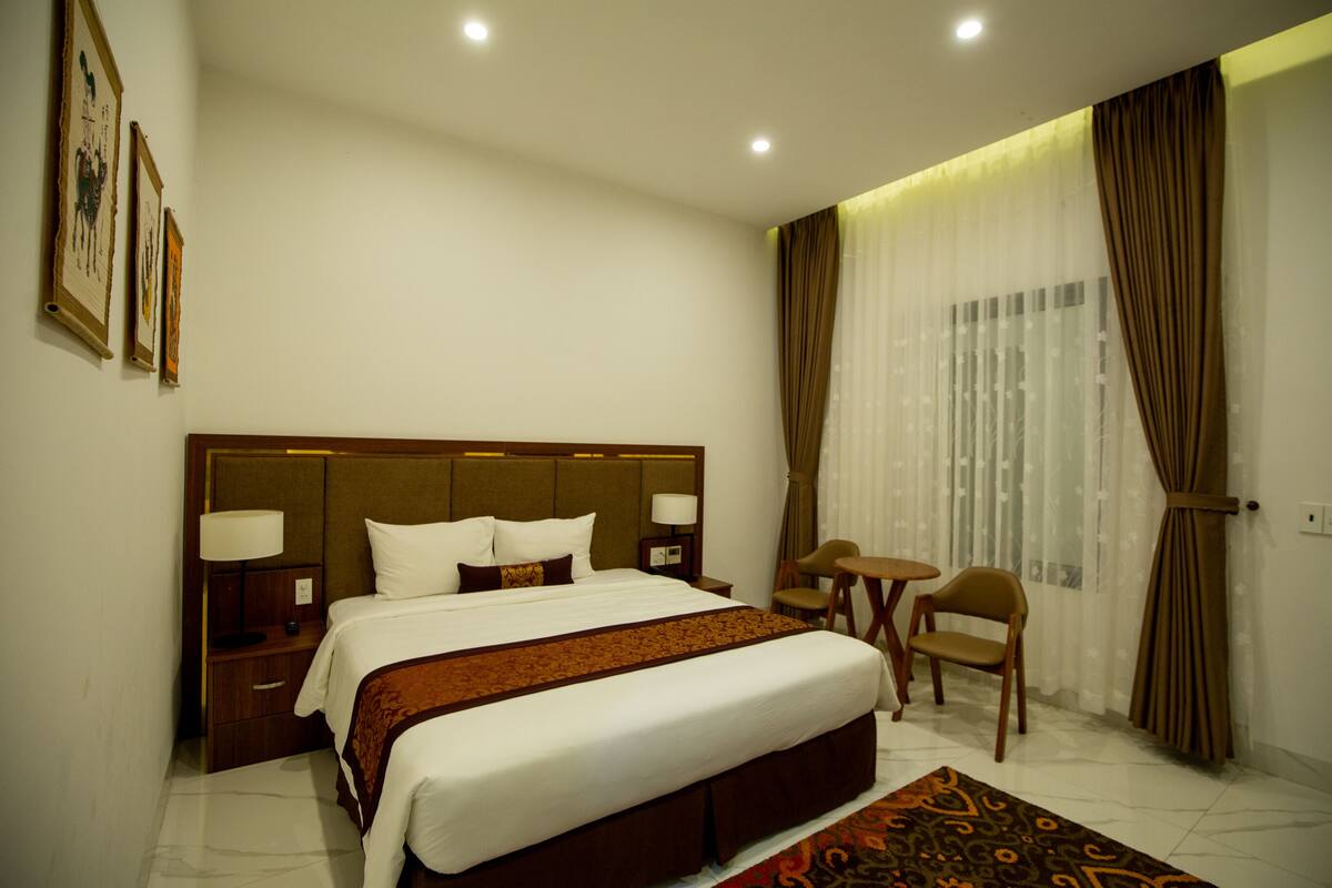A hotel room with a large bed, bedside tables, a small table and chair, and a window with curtains.