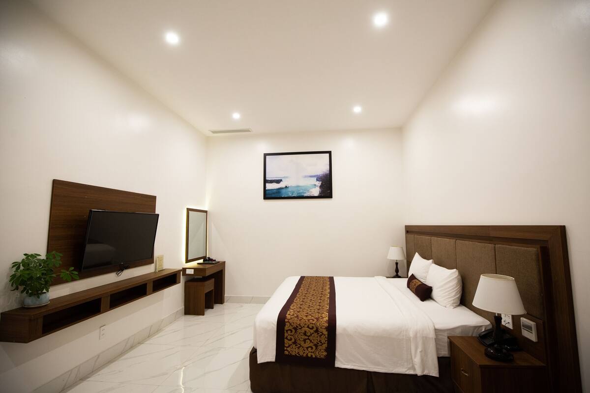 A hotel room with a bed, bedside table, lamp, TV, and a painting on the wall.