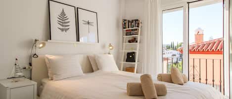 A neatly made bed with pillows and rolled towels, a bedside table with a lamp, a bookshelf with books, and a view of buildings through a balcony.