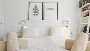 Un dormitorio minimalista con una cama grande, dos impresiones enmarcadas de motivos botánicos, lámparas de noche y una estantería.