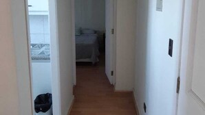 Een smalle gang met houten vloer die leidt naar een slaapkamer met een bed en een raam.