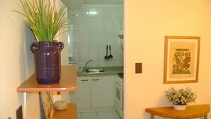 Een keuken met witte kastjes, een spoelbak en een fornuis. Op een plank staat een paarse vaas met groene planten, aan de muur hangt een ingelijst beeld en er staat een klein tafeltje met een plant.