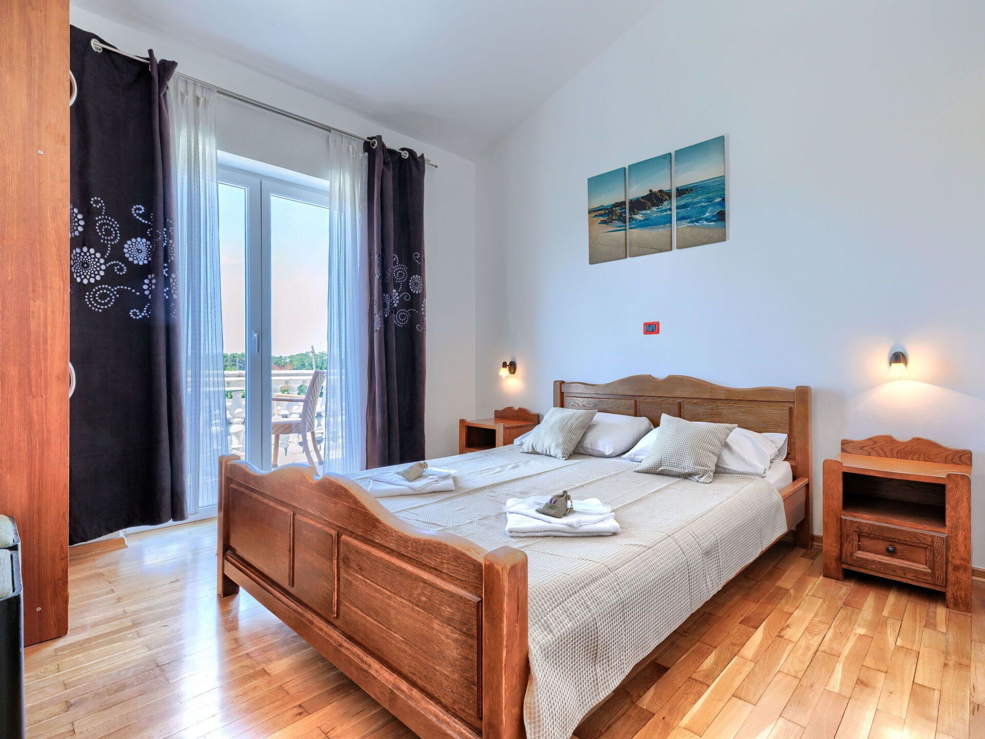 A bedroom with a wooden bed, two bedside tables, a view of the outdoors through a sliding door, and a painting on the wall.