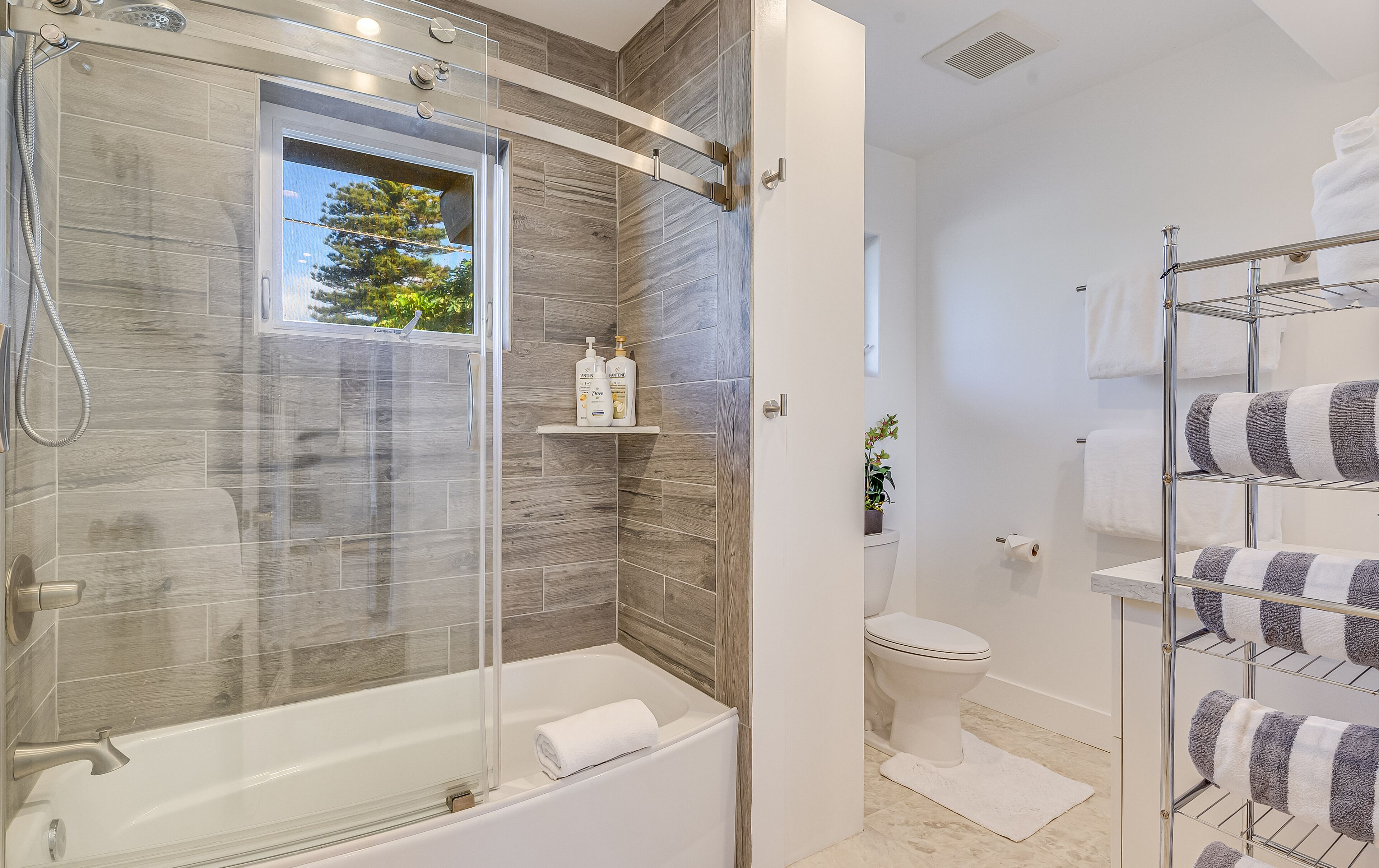 A modern bathroom with a glass-enclosed shower, a white bathtub, and a toilet. There are shelves with toiletries and a towel rack with striped towels.