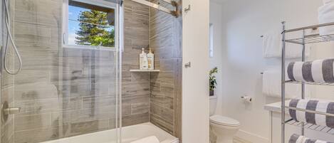 A modern bathroom with a glass-enclosed shower, a white bathtub, and a toilet. There are shelves with toiletries and a towel rack with striped towels.