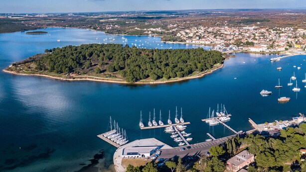 Vue aérienne d’une ville côtière avec un port de plaisance, une petite île couverte d’une forêt dense et un ciel dégagé.