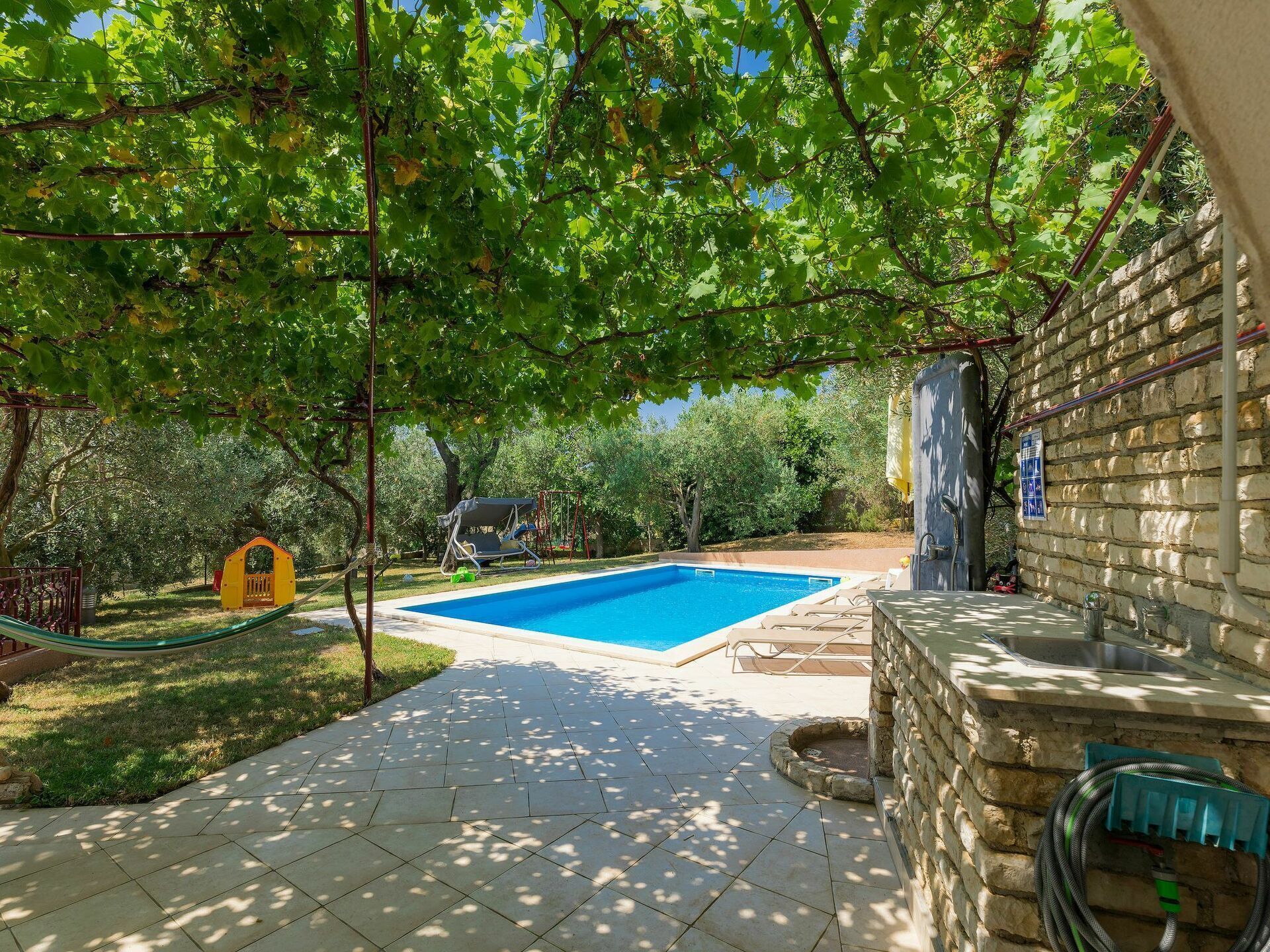 Un patio trasero con piscina, zona de juegos infantiles y un muro de piedra.