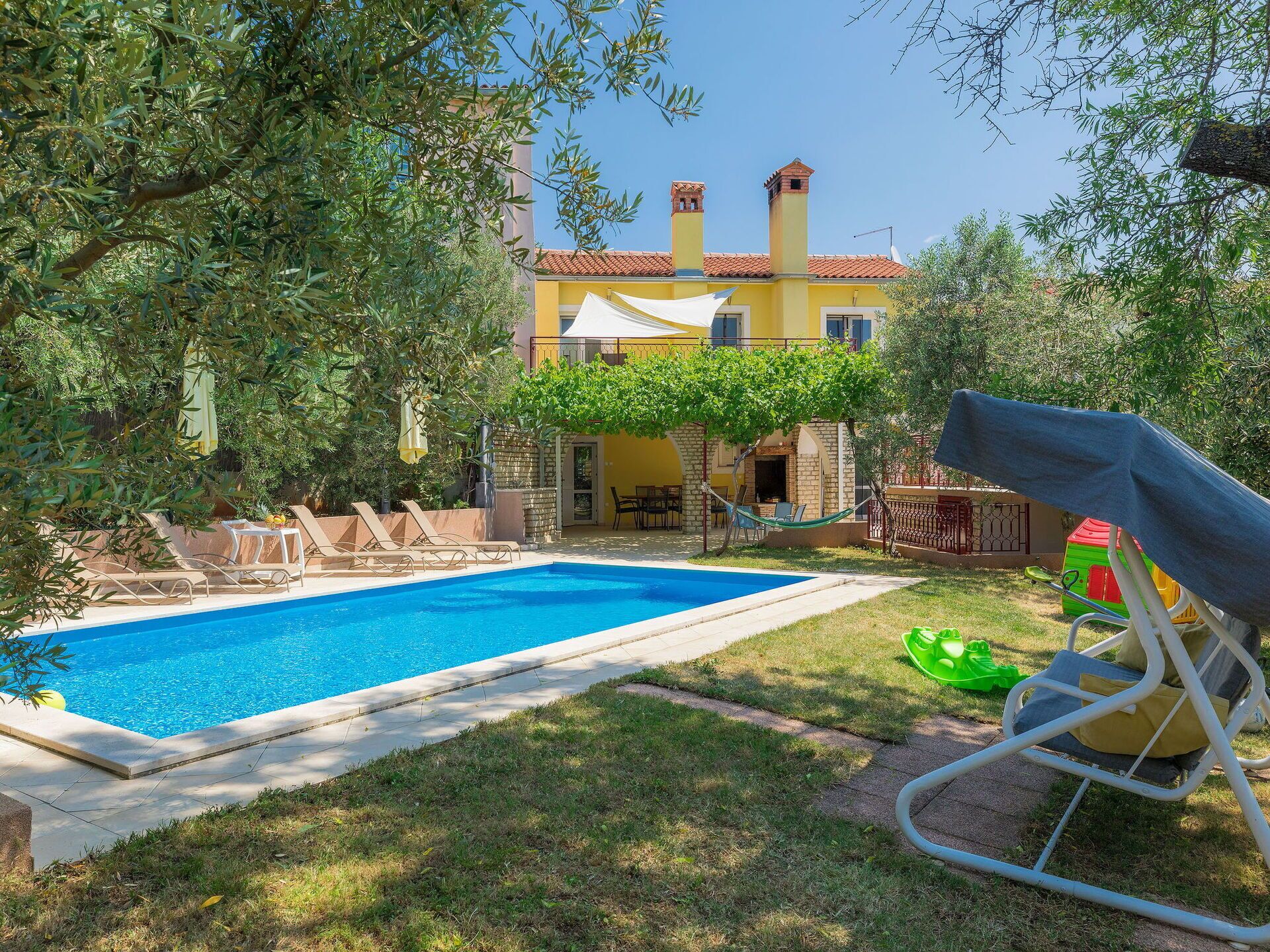 A swimming pool with lounge chairs, a green play structure, and a yellow building with a tiled roof in the background.