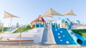 A playground with slides and climbing structures, surrounded by sand and greenery.