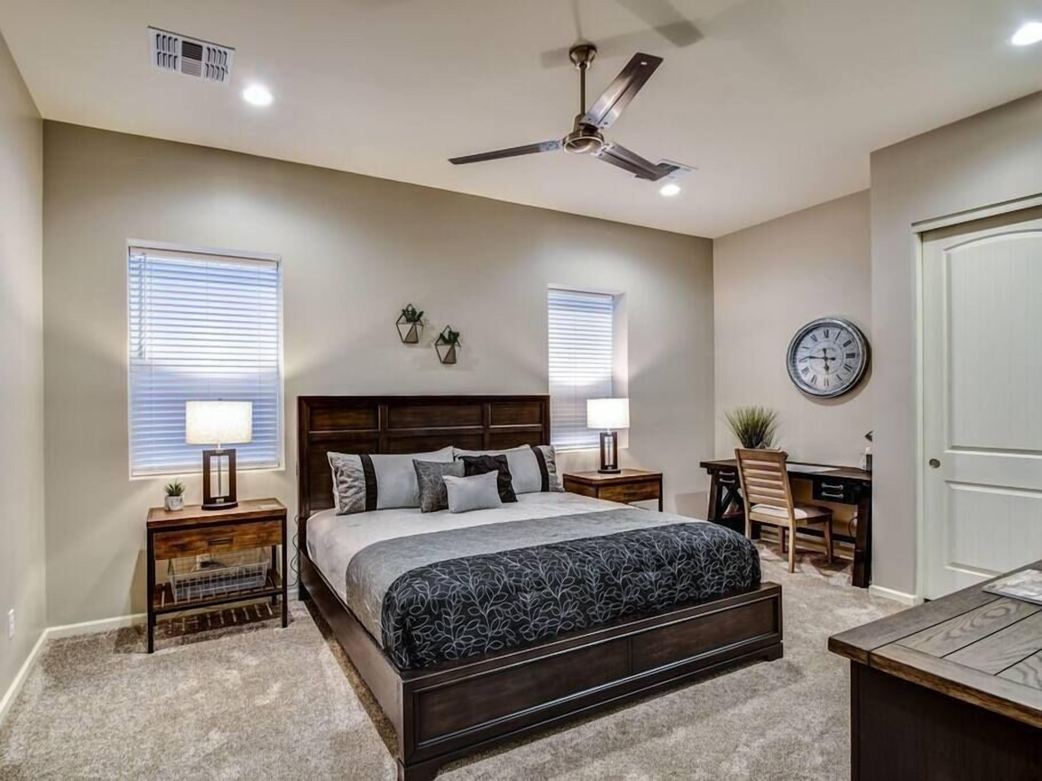 A bedroom with a bed, bedside tables, a desk, a chair, a ceiling fan, and a clock on the wall.