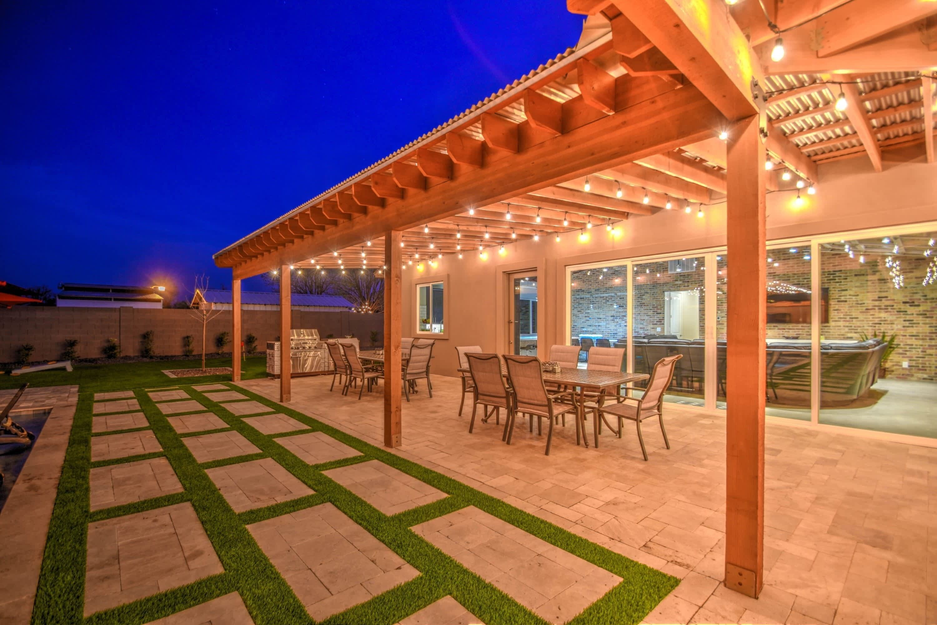 Área de terraço ao ar livre com pérgola, luzes de cordão e uma mesa de jantar com cadeiras.