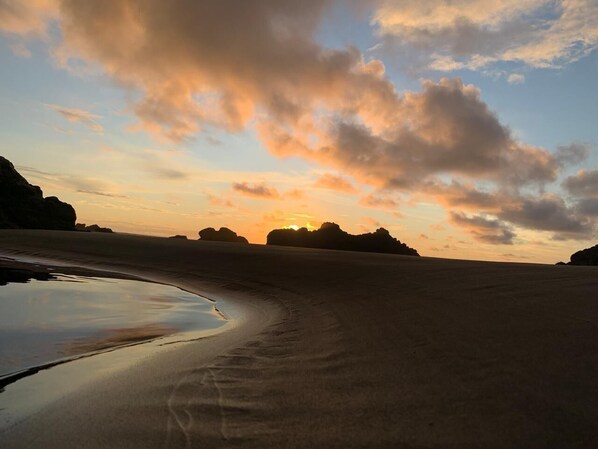 Escena de playa con una masa de agua tranquila que refleja el cielo, rodeada de arena y formaciones rocosas.