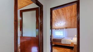 Una habitación con piso de madera, cama con ropa blanca y ventana con cortinas traslúcidas.
