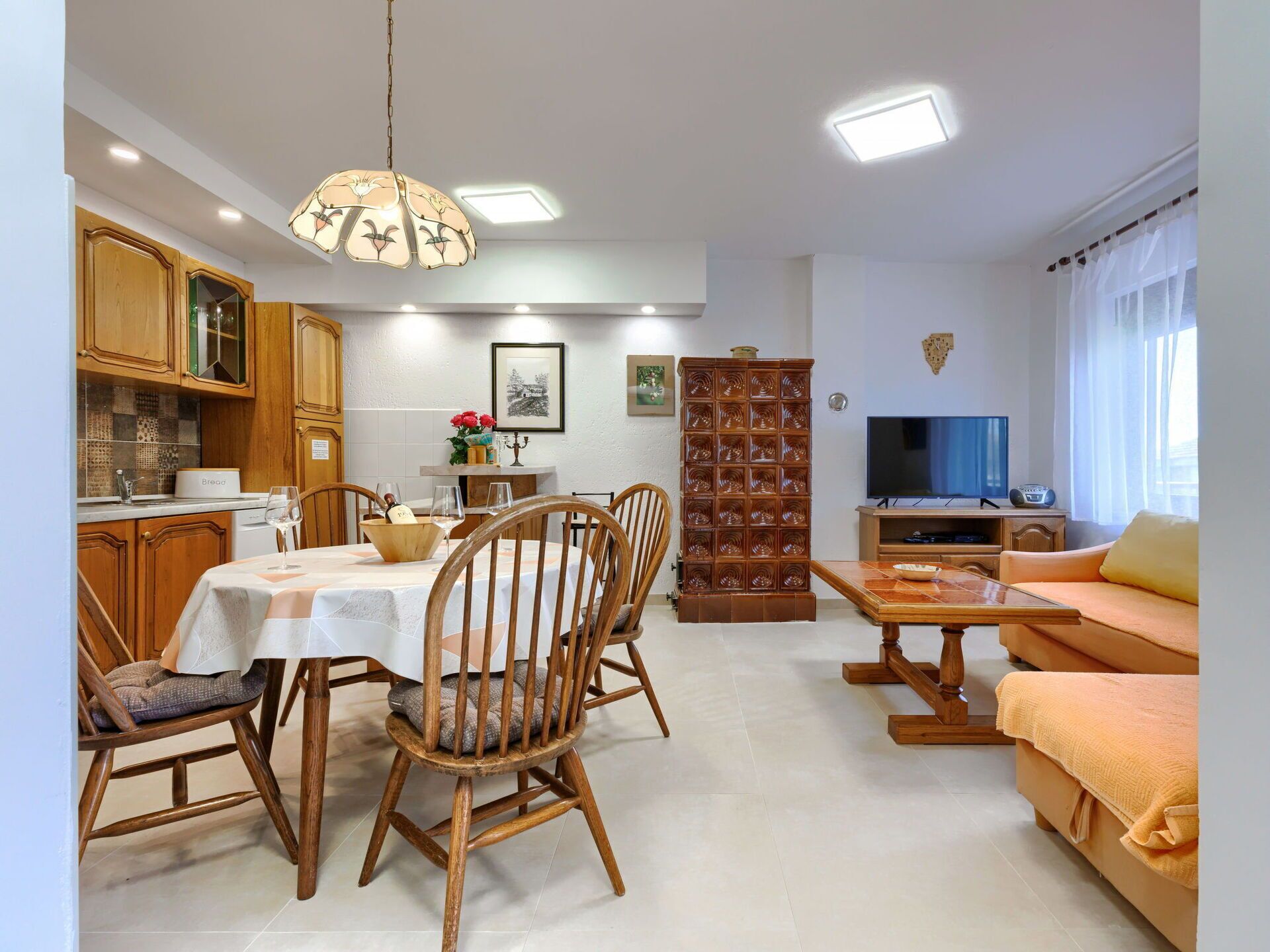 A dining area with wooden chairs and a table, a living room with a sofa and a television, and a kitchen with wooden cabinets and a BREAD machine.