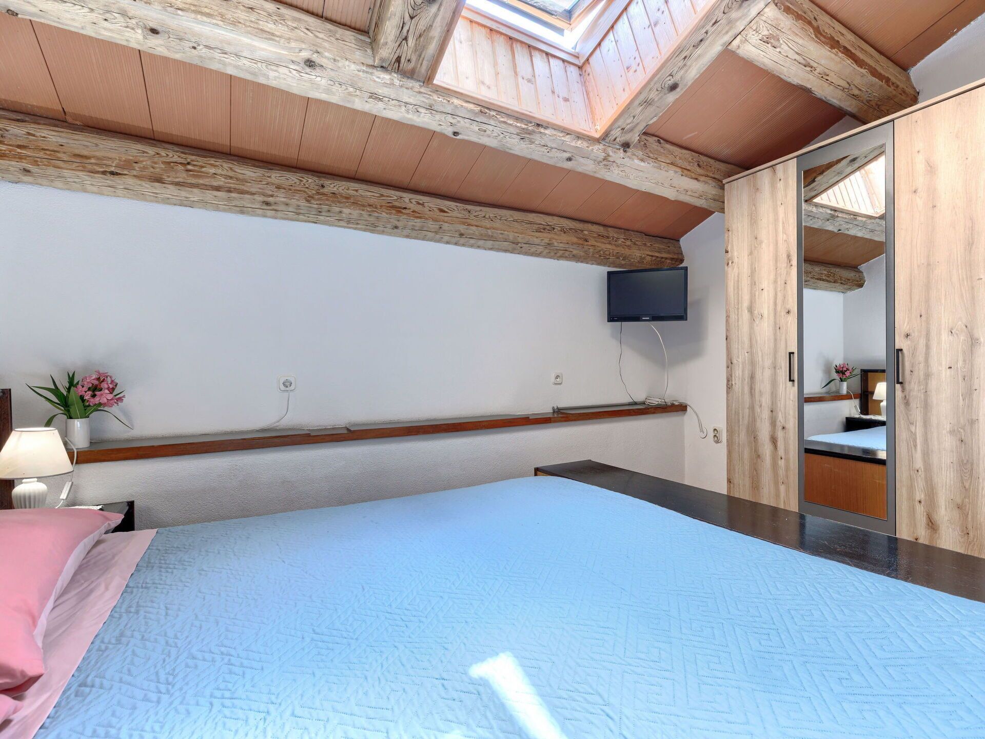 Une chambre à coucher avec un lit, un téléviseur fixé au mur, un plafond en bois et un puits de lumière.