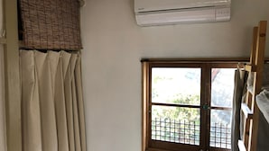 Una habitación con ventana protegida por una reja metálica, cortinas color beige, una escalera de madera y un equipo de aire acondicionado en la pared.